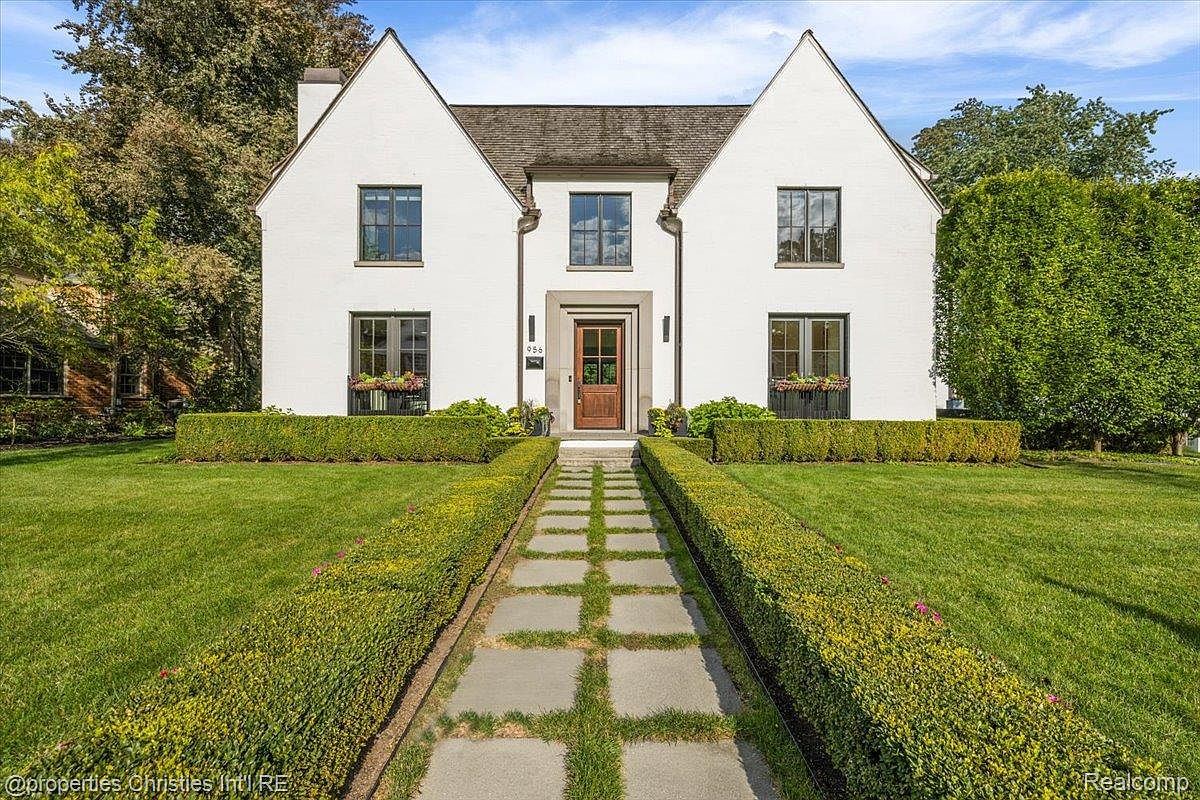 This is a front view of a stately, symmetrical white house with a well-manicured lawn and garden. A stone pathway leads to the front door, flanked by neatly trimmed hedges. The house features multiple windows with dark frames and a classic architectural style, creating an impression of elegance and curb appeal.