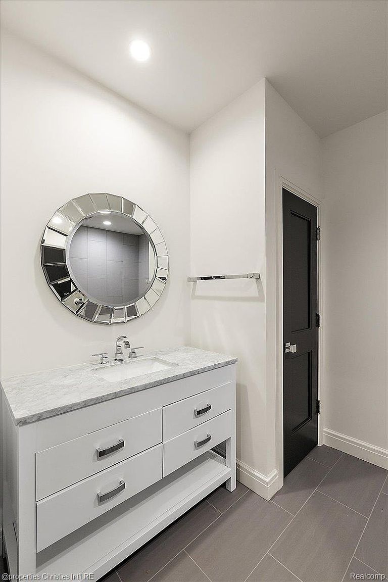 This is a well-lit bathroom featuring a white vanity with a marble countertop and chrome fixtures. A round, decorative mirror hangs above the sink, and a dark door is visible to the right. The floor is tiled in a neutral gray, creating a clean and modern aesthetic.