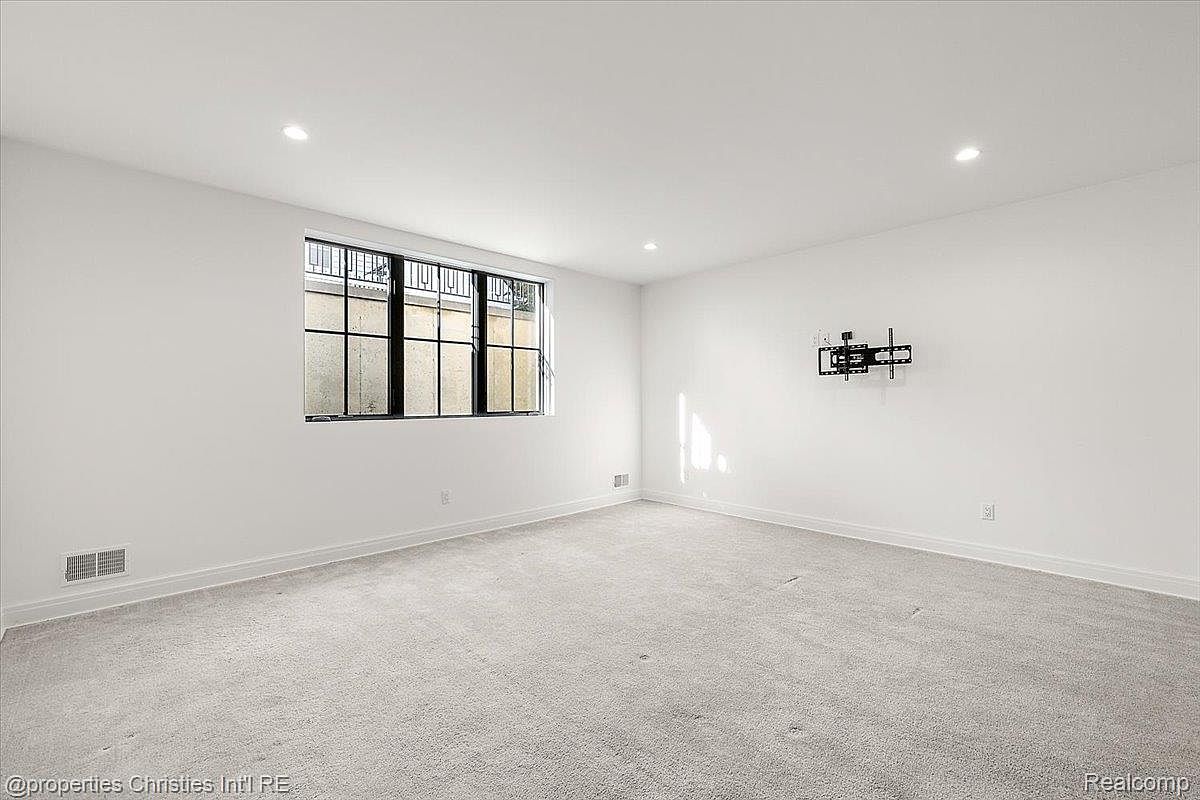 The image showcases a spacious basement area with neutral-toned carpeting and white walls. A window provides natural light, and a wall-mounted TV bracket suggests entertainment possibilities. The room appears clean and well-maintained, offering a blank canvas for various uses.