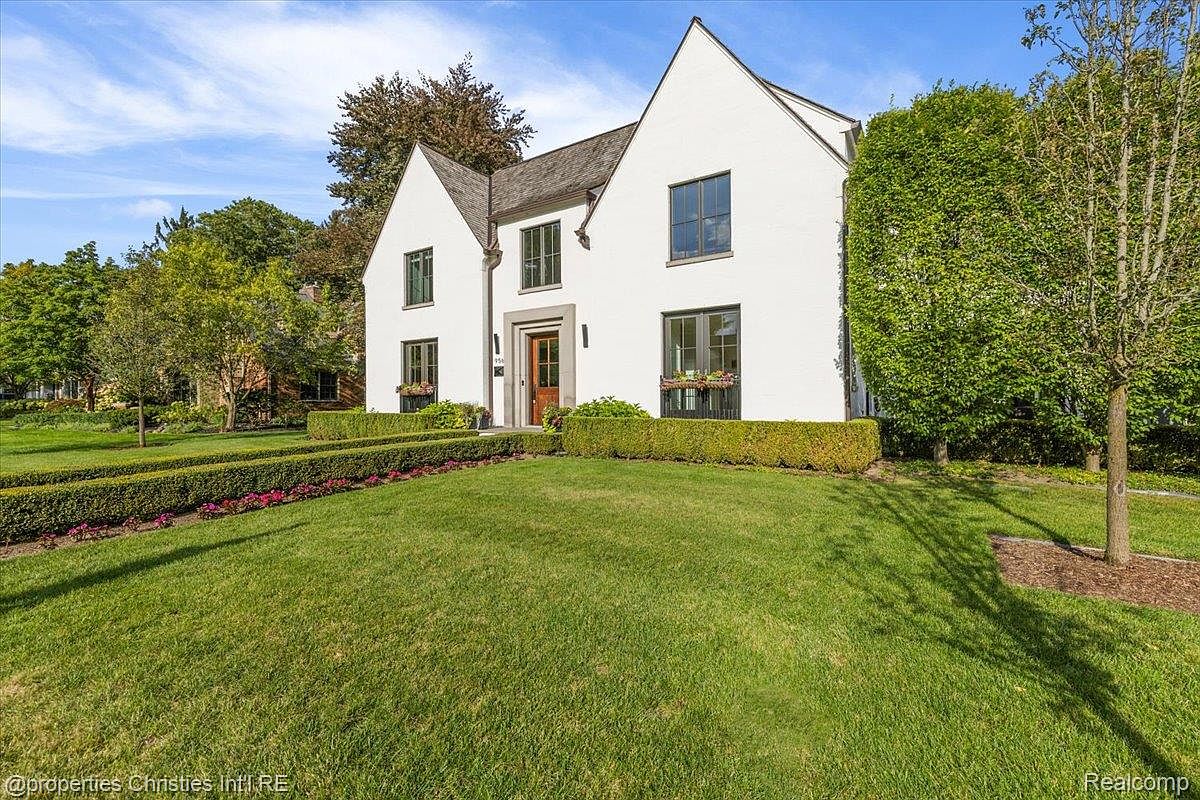 This is a front exterior view of a two-story white house with a well-manicured lawn. The house features a symmetrical design with a dark roof, dark-framed windows, and a central entryway. The landscaping includes trimmed hedges and a lush green lawn, creating a welcoming and elegant curb appeal.