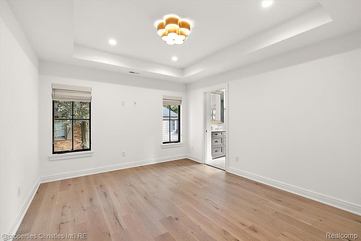 This is a bright and spacious primary bedroom featuring hardwood floors, white walls, and two windows that provide natural light. The room has a modern recessed ceiling with a decorative light fixture. An open doorway leads to an en-suite bathroom, adding to the convenience and appeal of the space.