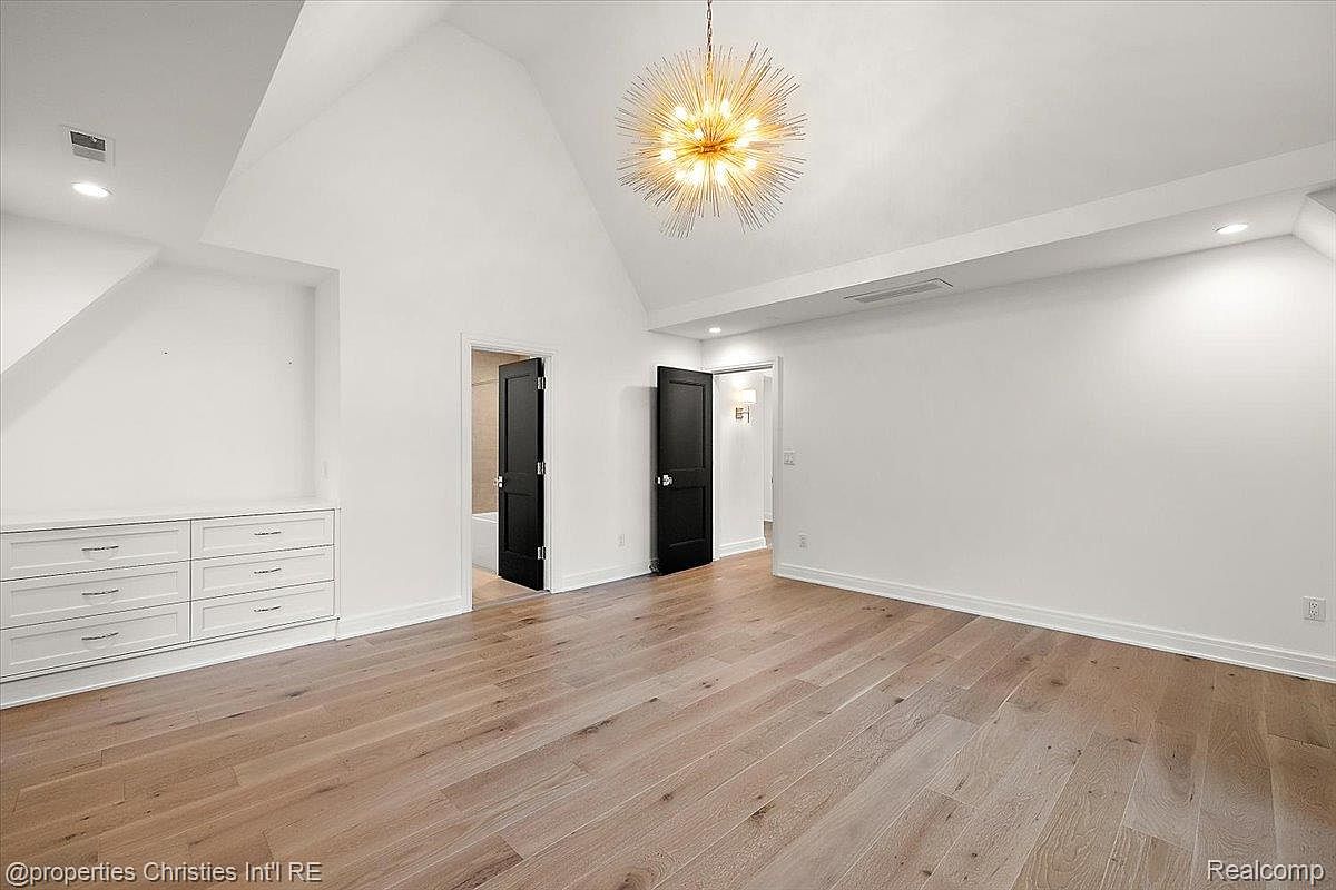 This is a spacious primary bedroom featuring light hardwood floors and white walls. A modern chandelier hangs from the vaulted ceiling, adding a touch of elegance. Built-in drawers provide ample storage, and two dark doors lead to other areas of the house, creating a seamless flow.