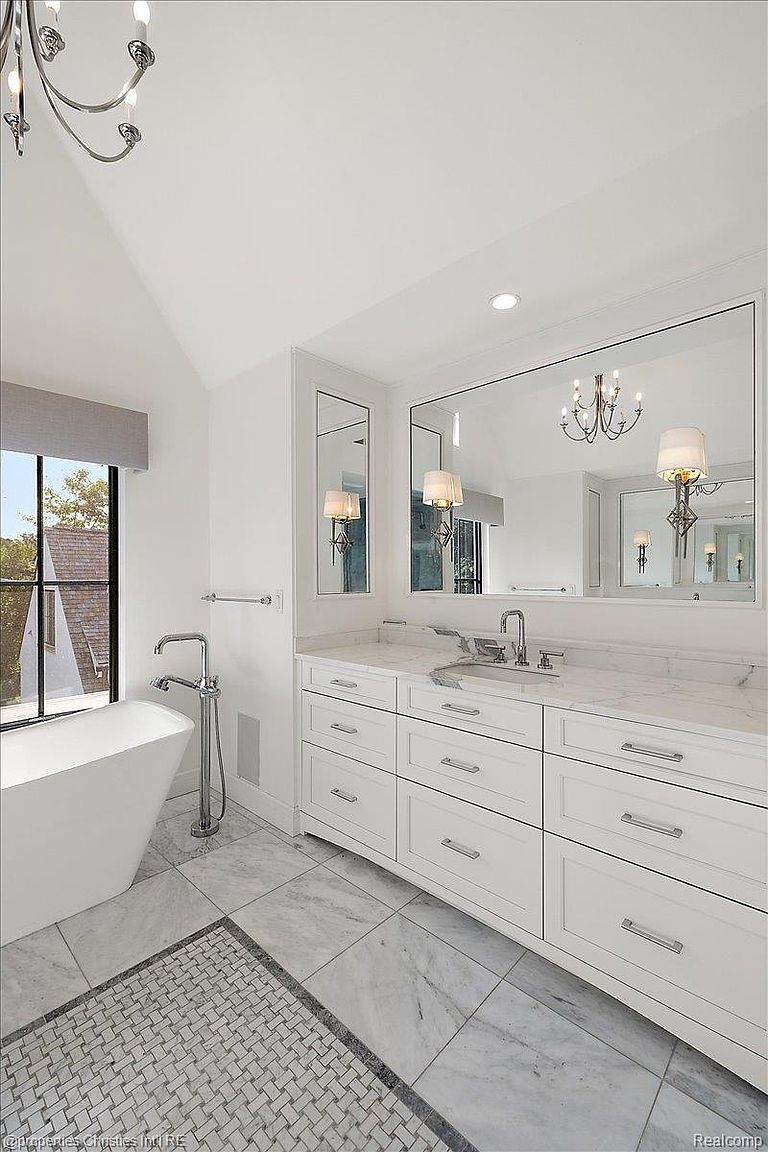 This is a bright and elegant primary bathroom featuring a double vanity with white cabinetry and marble countertops. A large mirror reflects the space, enhancing the sense of openness, while a freestanding bathtub sits near a window, offering a relaxing view. The flooring is marble tile, complemented by a decorative rug.