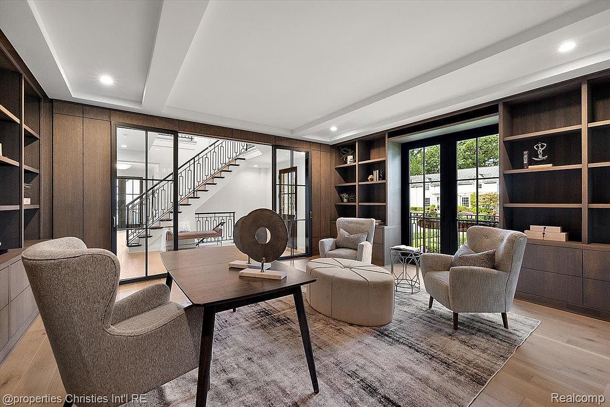 This is an interior shot of a sophisticated home office or study. The room features built-in shelving and cabinetry, a large desk with a decorative sculpture, and comfortable armchairs with an ottoman. A glass-enclosed staircase is visible in the background, adding to the open and airy feel of the space.
