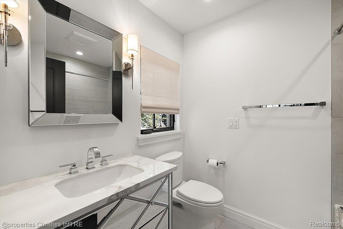 This is a well-lit guest bathroom featuring a modern vanity with a white marble countertop and chrome fixtures. A large mirror hangs above the sink, reflecting the clean lines of the space. The toilet is positioned next to a window with a neutral-toned blind, and a chrome towel bar adds a touch of elegance to the white walls.