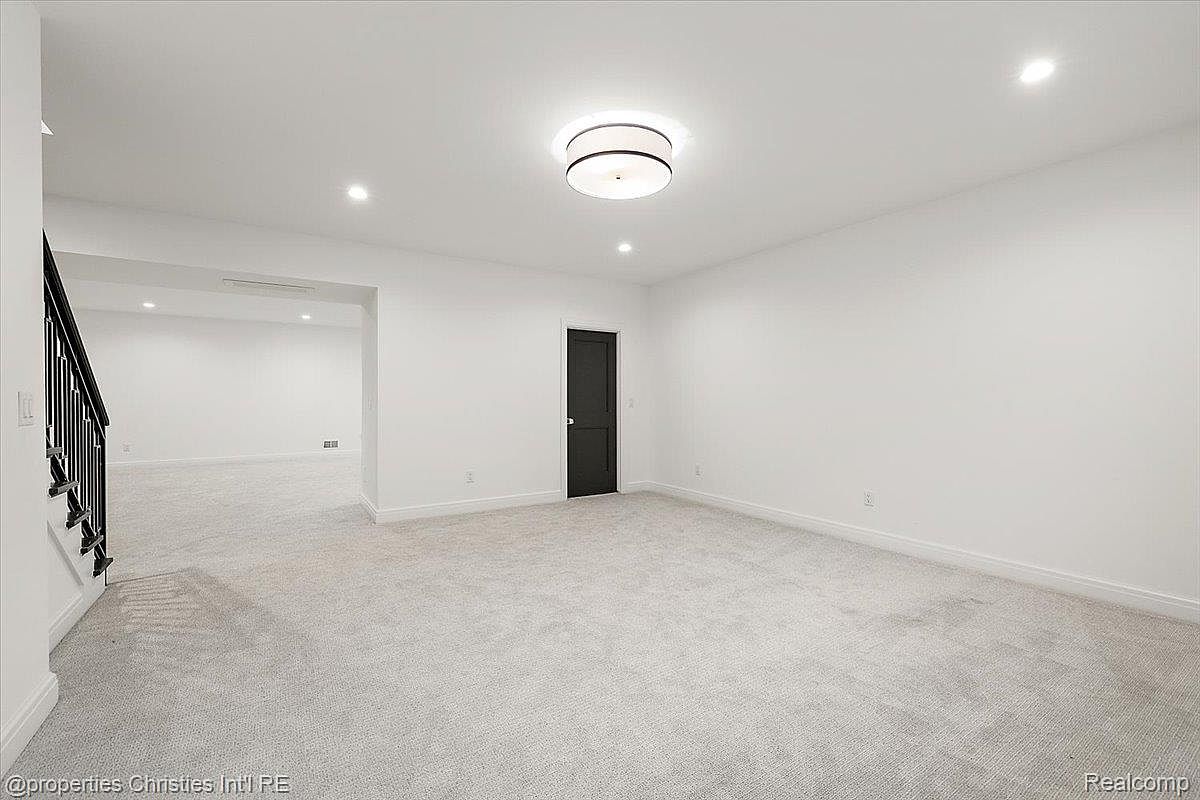This is a spacious, well-lit basement featuring neutral-toned carpeting and white walls. A modern ceiling light fixture provides ample illumination, and a dark-colored door adds a touch of contrast. The open layout suggests versatility for various uses, such as a recreation room or home theater.