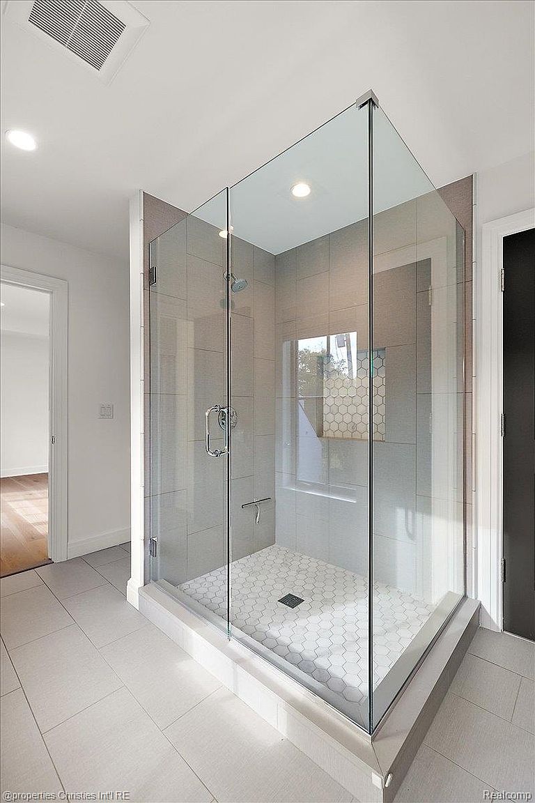 This is a well-lit primary bathroom featuring a modern, frameless glass shower enclosure with gray tiled walls and a white hexagonal tiled floor. The shower includes a rainfall showerhead and sleek hardware. The overall impression is clean, contemporary, and luxurious, highlighting the updated features of the bathroom.
