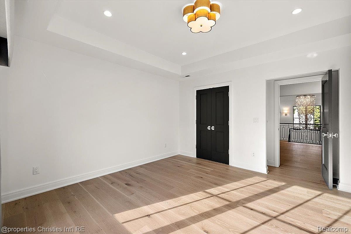 This is an interior shot of a bedroom featuring light hardwood floors and white walls. A black double door is centered on one wall, and an open doorway to another room is visible on the right. The room is illuminated by a modern, flower-shaped ceiling light fixture and natural light streaming through the doorway.