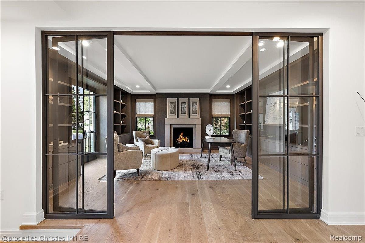 This interior shot showcases a sophisticated living room with a fireplace, built-in bookshelves, and elegant furnishings. The room is framed by dark-framed glass doors, offering a glimpse into the space and highlighting the open layout. The hardwood flooring and neutral color palette create a warm and inviting atmosphere.