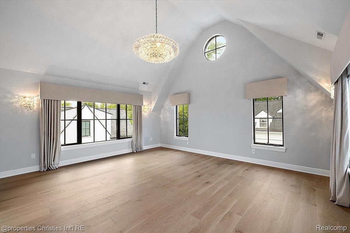 This is a spacious primary bedroom featuring light hardwood floors and a high, vaulted ceiling. Large windows provide ample natural light, complemented by elegant wall sconces and a decorative chandelier. The room exudes a sense of calm and luxury, perfect for relaxation.