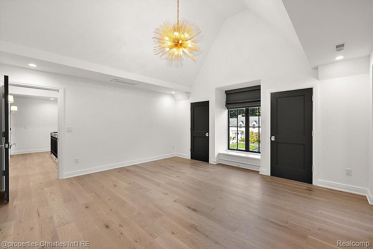 This is a spacious primary bedroom featuring light hardwood floors and white walls, creating a bright and airy atmosphere. The room includes two dark-colored doors, a window with dark trim and blinds, and a modern chandelier. The perspective is from the doorway, showcasing the room's size and layout.