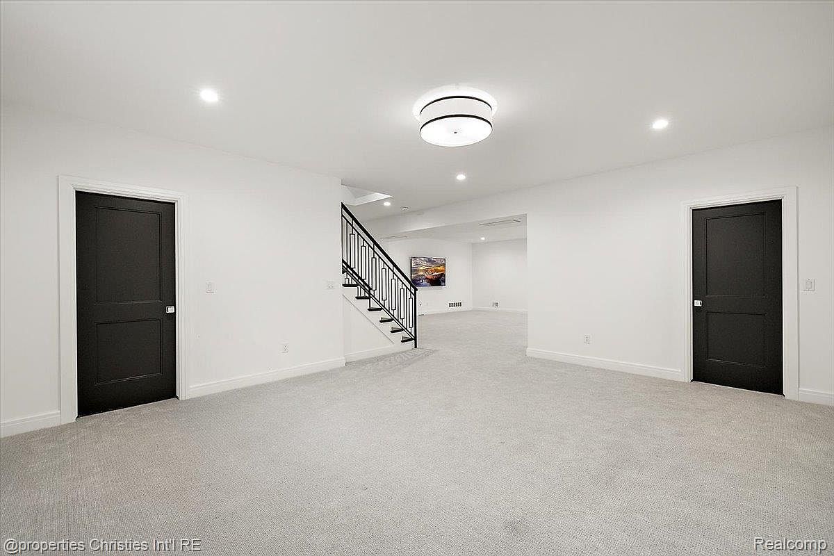 This is a spacious basement featuring neutral gray carpeting and white walls, creating a bright and open feel. Two dark-colored doors are positioned on opposite sides of the room, and a staircase with black railings leads to the upper level. A modern ceiling light fixture adds a touch of style to the room.