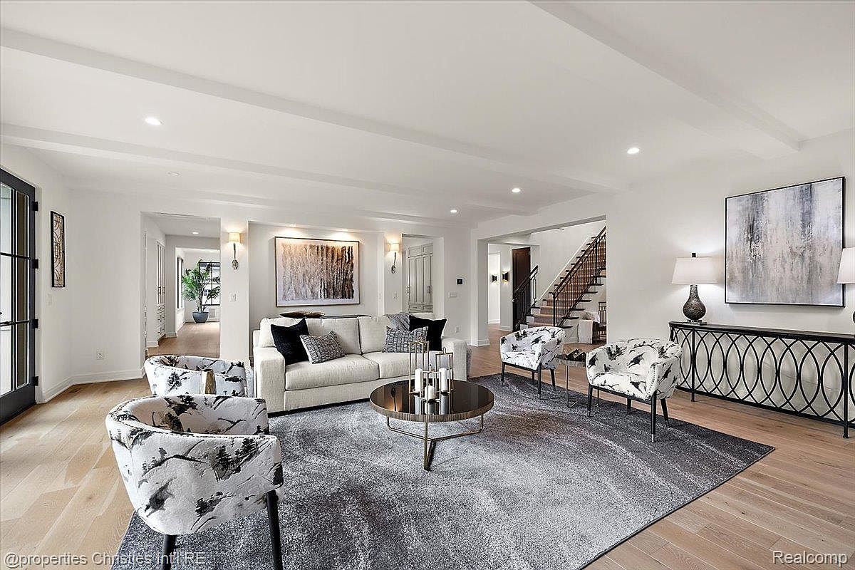 This is an interior shot of a living room featuring a neutral color palette with hardwood floors and a large area rug. The room is furnished with a light-colored sofa, patterned armchairs, and a round coffee table. Artwork adorns the walls, and a console table with lamps sits against one wall, creating a sophisticated and inviting atmosphere.