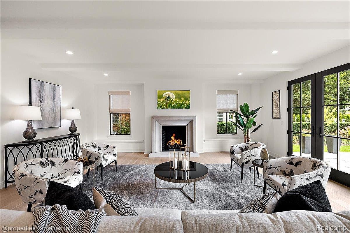 This is a bright and spacious living room featuring a fireplace, large windows, and stylish furnishings. The room is decorated in a neutral color palette with pops of black and white in the patterned armchairs and throw pillows. A round coffee table sits in the center of the room on top of a gray rug, and a console table with lamps is placed against one wall. The perspective is from the sofa, giving a sense of being in the room.