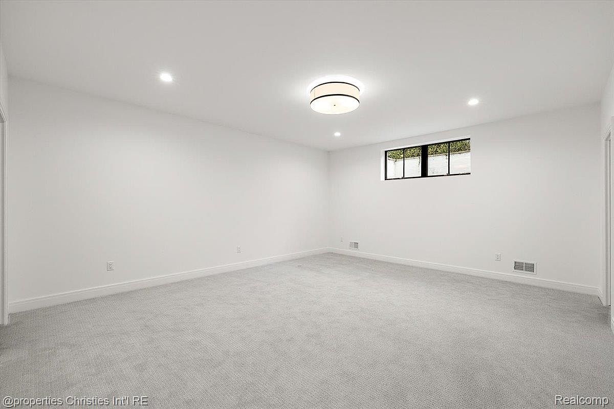 The image showcases a spacious, finished basement with neutral gray carpeting and white walls, creating a bright and open feel. A small window provides natural light, and recessed lighting and a modern ceiling fixture illuminate the room. The space appears clean and versatile, suitable for various uses such as a recreation room or home theater.