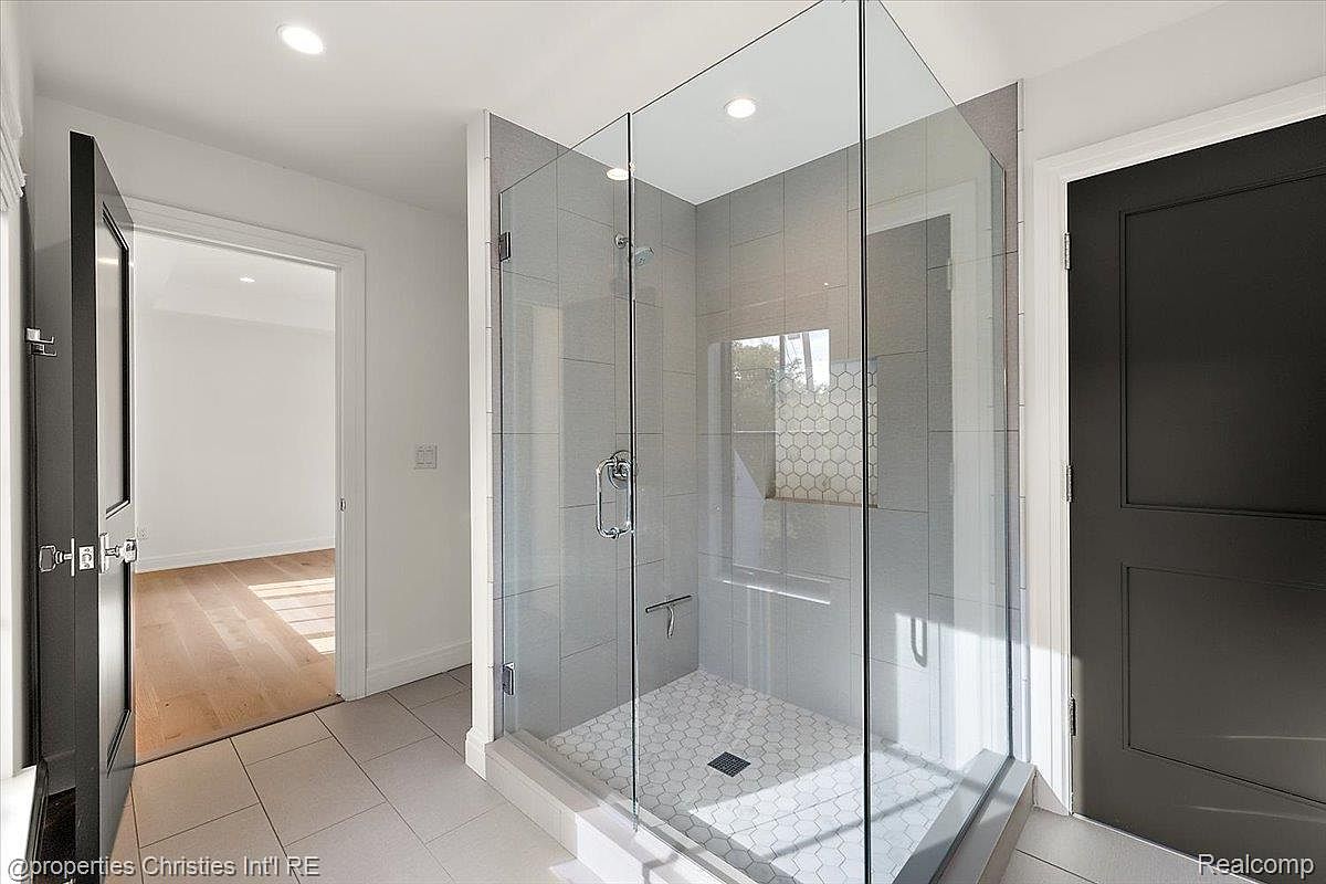 This is a well-lit primary bathroom featuring a modern glass-enclosed shower with gray tiling and a white hexagonal tile floor. The shower has a sleek design with a chrome shower head and handle. A dark-colored door is visible to the right, and an open doorway leads to another room with hardwood floors, creating a sense of spaciousness and flow.