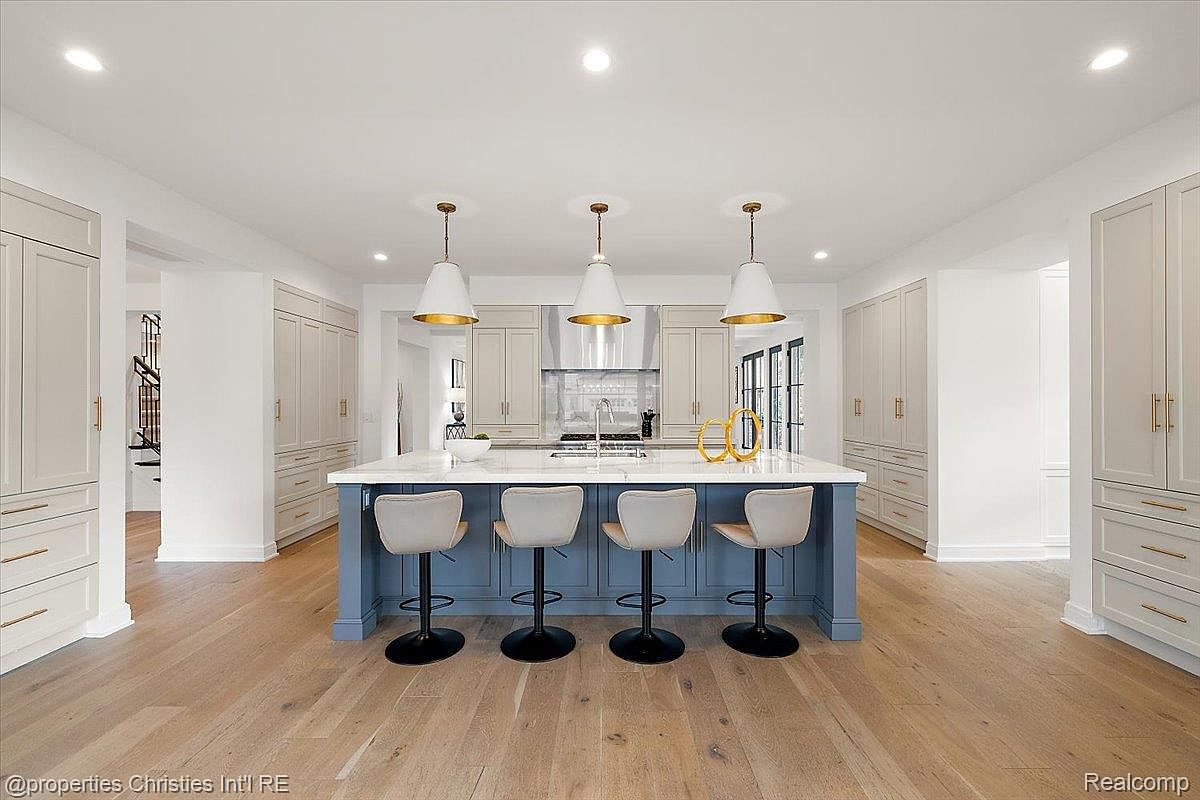 This is a bright and spacious kitchen featuring a large island with a blue base and white countertop, complemented by four bar stools. The kitchen is surrounded by light gray cabinetry with gold hardware, and three pendant lights hang above the island, adding a touch of elegance. The hardwood flooring adds warmth to the space, creating an inviting and modern atmosphere.