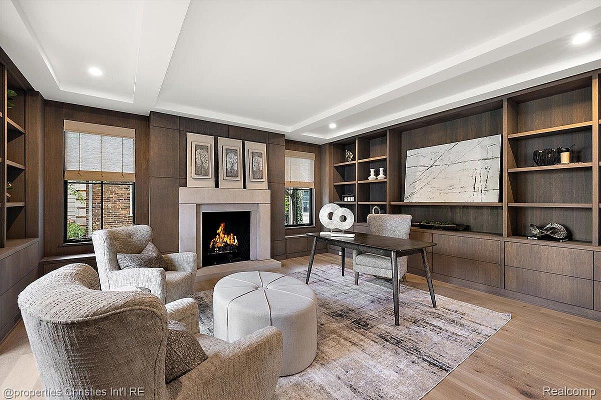 This is an interior shot of a sophisticated home office or study. The room features dark wood built-in shelving, a stone fireplace, and comfortable seating including armchairs and an ottoman. A desk with a modern sculpture sits on a patterned rug, creating a professional yet inviting atmosphere.