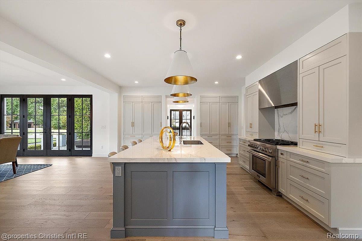 This is a bright and spacious kitchen featuring a large center island with a marble countertop and gray base. The kitchen is equipped with stainless steel appliances and ample white cabinetry, complemented by gold hardware. The open floor plan leads to a living area with large glass doors, enhancing the sense of space and light.