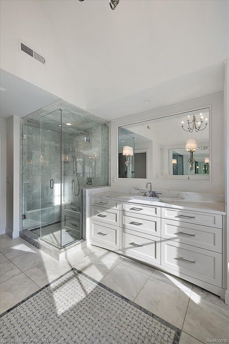 This is a bright and luxurious primary bathroom featuring a glass-enclosed shower with marble walls and a built-in bench. A double vanity with white cabinetry and marble countertops is complemented by a large mirror reflecting a chandelier, enhancing the sense of space and elegance. The floor is tiled with a decorative mosaic pattern, adding a touch of sophistication.