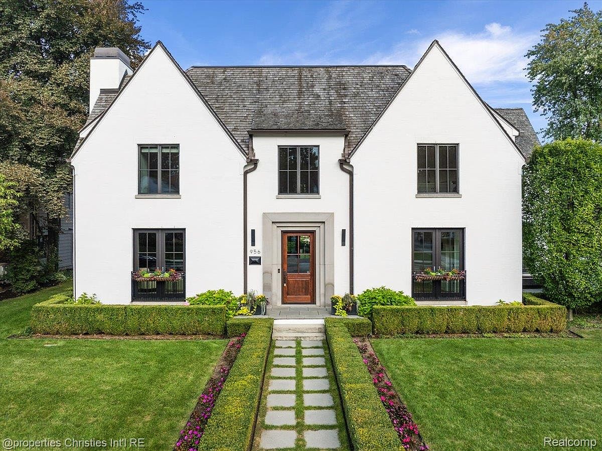 This is a front view of a charming two-story house with a symmetrical design. The exterior is painted white, and the roof is a dark gray. The house features multiple windows with black frames and a well-manicured lawn with hedges and a stone pathway leading to the front door, creating a welcoming and elegant curb appeal.