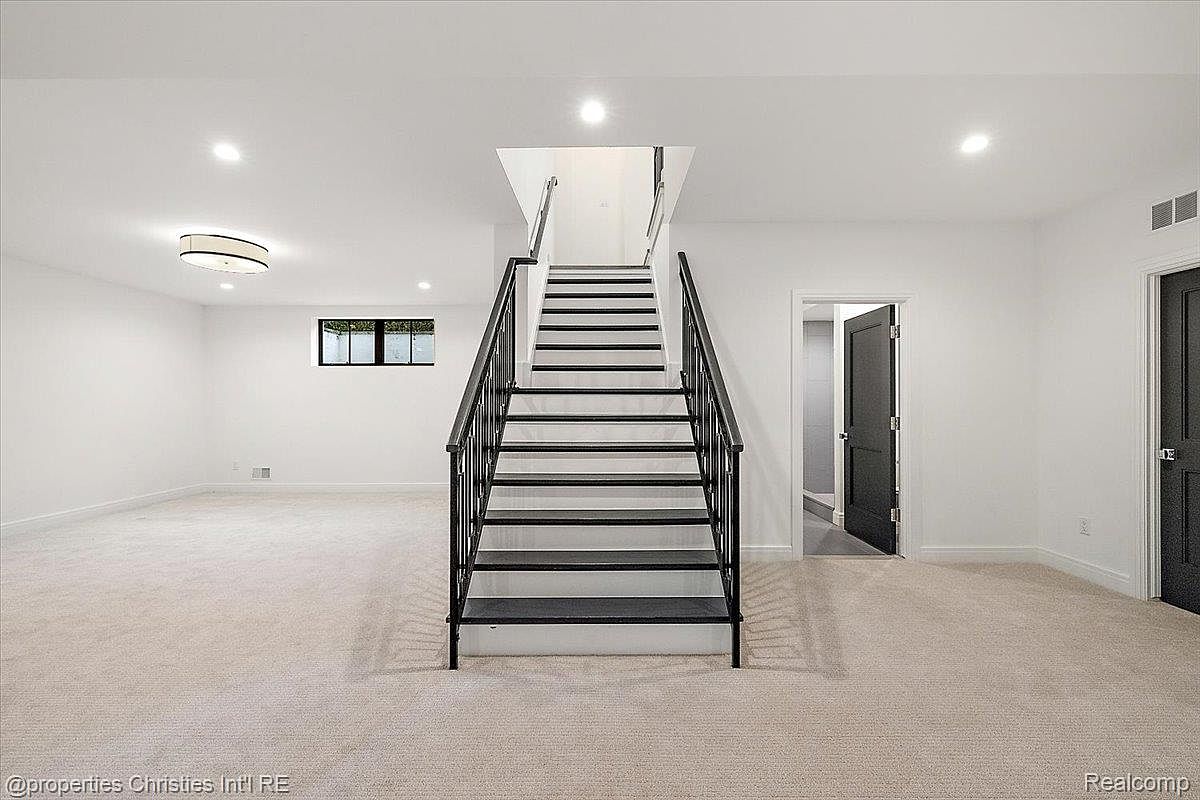 This is a spacious basement featuring a staircase leading to the upper level. The room is well-lit with recessed lighting and includes a window for natural light. The walls are painted white, and the floor is covered with light-colored carpet, creating a clean and inviting atmosphere.