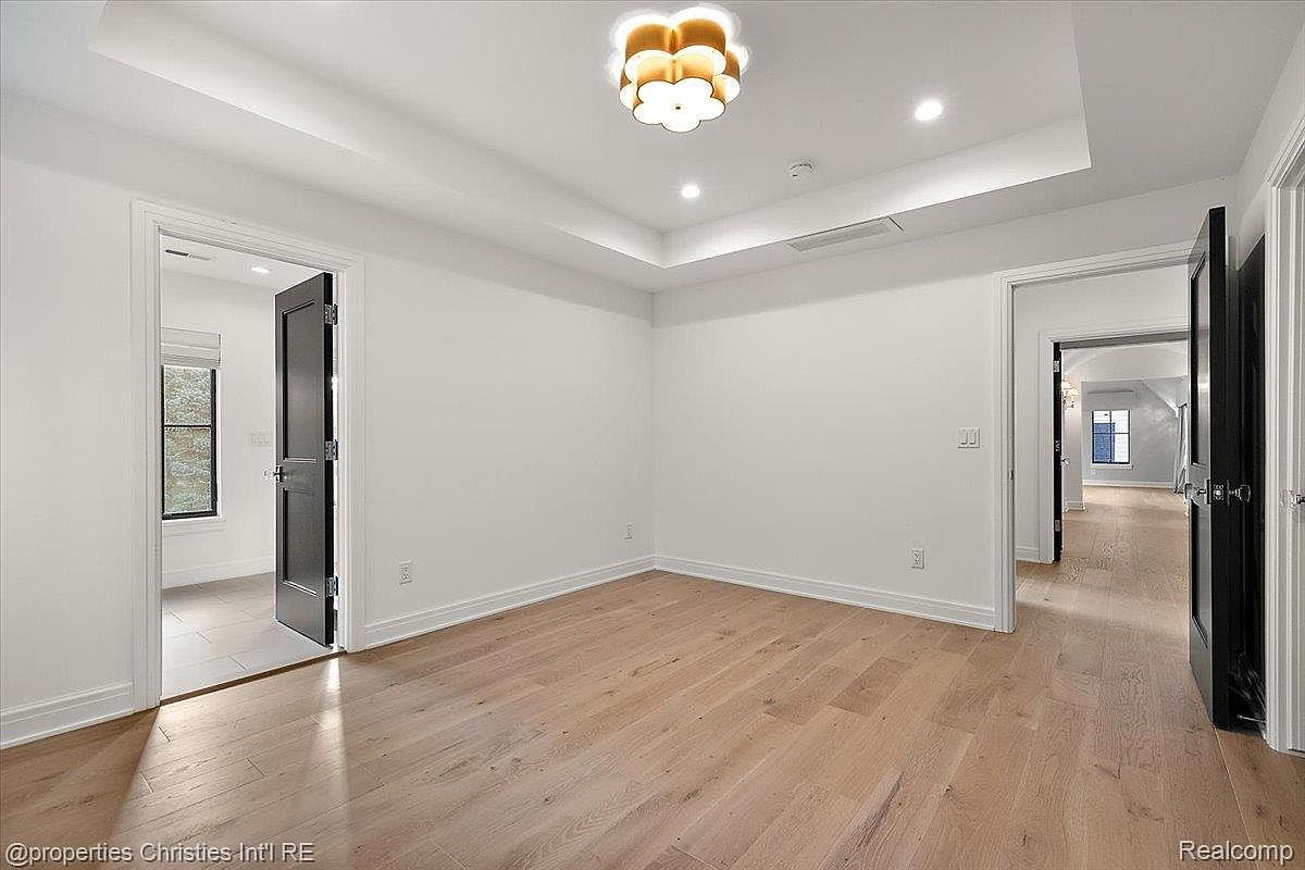 This is an interior shot of a bedroom featuring light hardwood floors and white walls, creating a bright and airy atmosphere. The room is illuminated by a modern, flower-shaped ceiling light fixture and recessed lighting. Two doorways, each with a dark-colored door, lead to other areas of the house, enhancing the sense of spaciousness and connectivity.