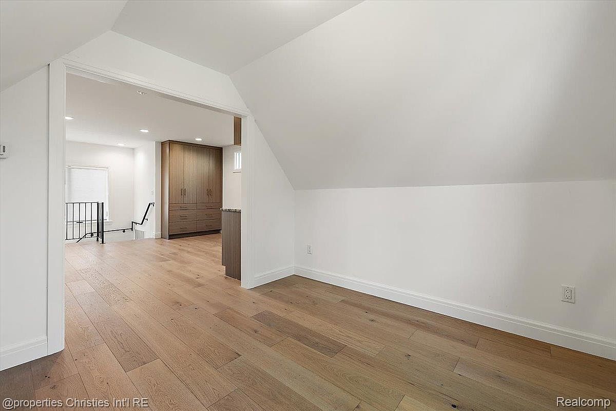 The image showcases a bright and airy attic space with hardwood flooring and white walls, creating a clean and modern aesthetic. The room features a distinctive sloped ceiling and an open doorway leading to another area, possibly a hallway or landing. The space appears versatile and well-maintained, suitable for various uses such as a bedroom, office, or storage area.