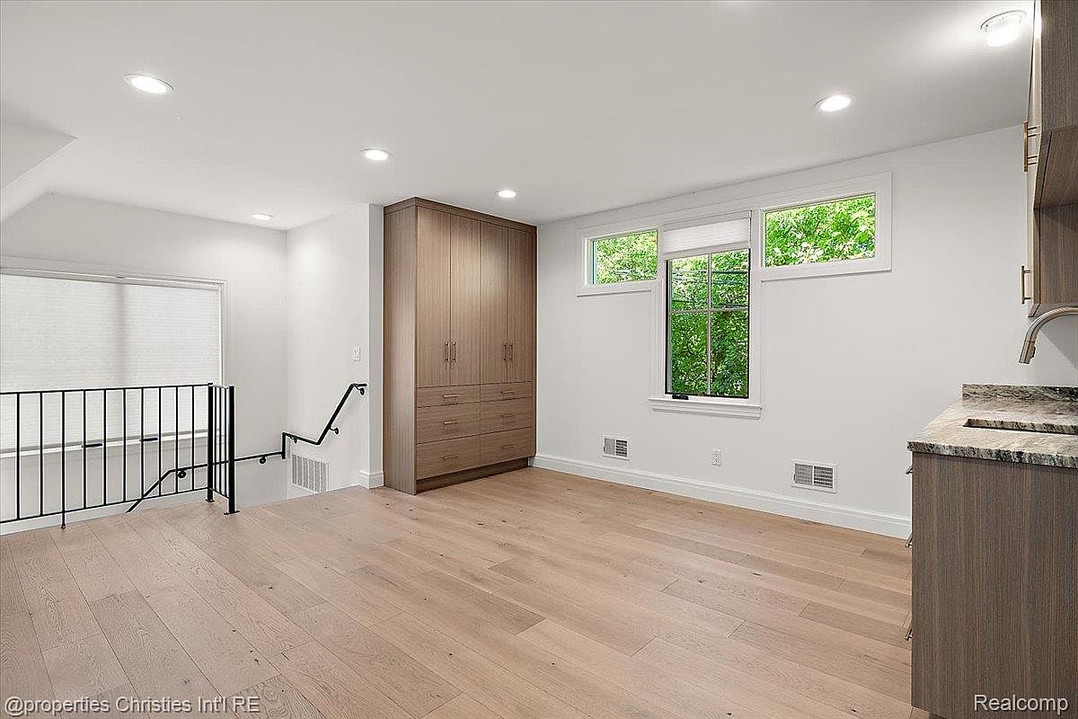 This is an interior shot of a living room featuring light hardwood floors and white walls. A built-in wooden cabinet provides storage, while natural light streams in through the windows. The room also includes a staircase with a black railing, adding architectural interest.