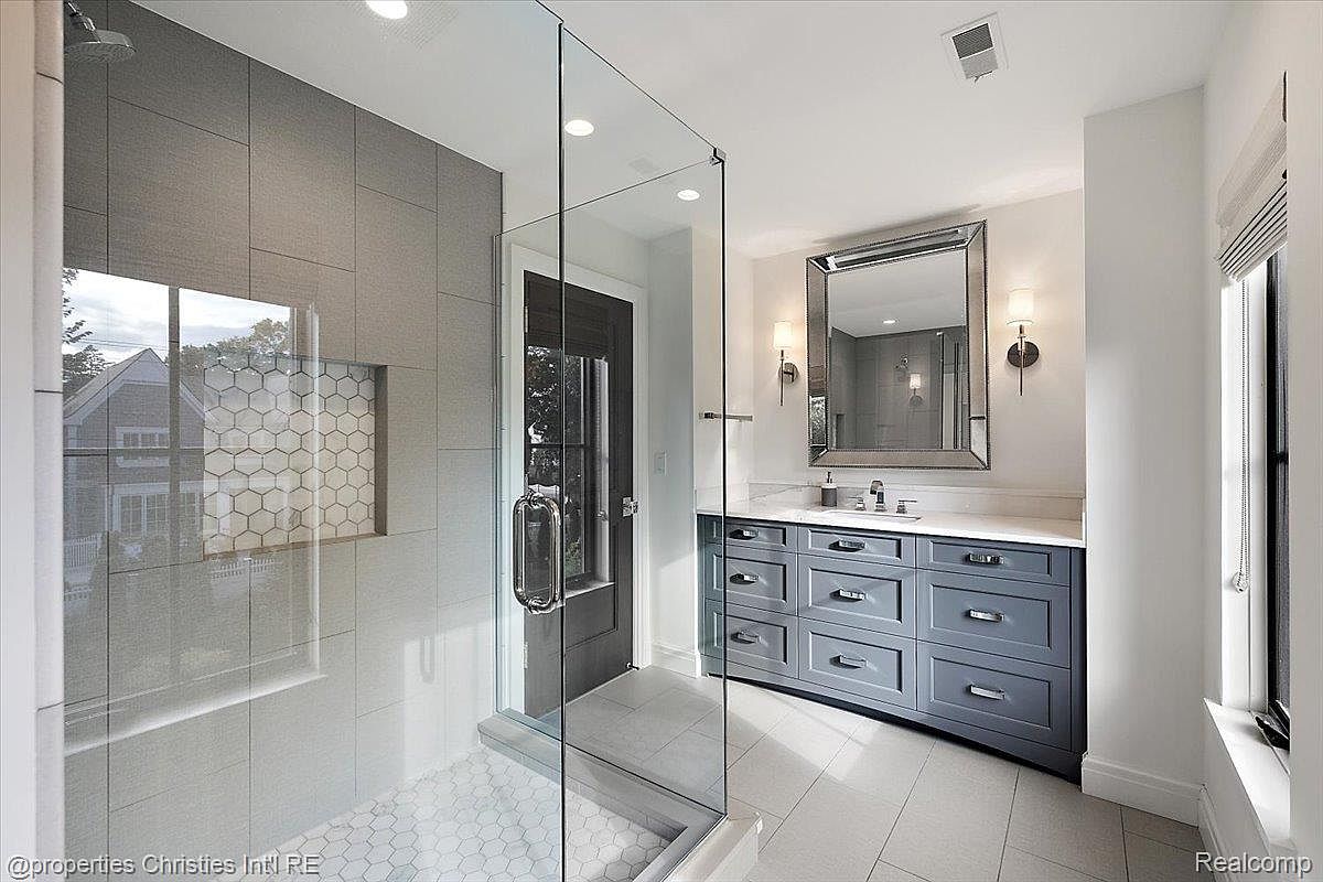 This is a primary bathroom featuring a glass-enclosed shower with gray and white tiling, a gray vanity with a white countertop, and a large mirror with sconces. The bathroom has a modern and clean design with neutral colors, creating a spa-like atmosphere. The flooring is light-colored tile, and there is a window providing natural light.