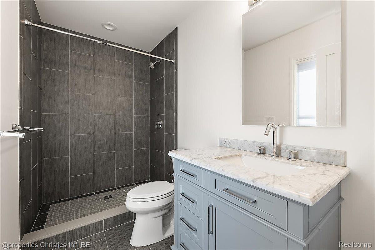 This is a well-lit bathroom featuring a modern design. The shower area is tiled in a dark gray, contrasting with the white walls and light gray vanity. The vanity has a marble countertop and silver hardware, and a toilet is positioned next to the shower, creating a functional and stylish space.