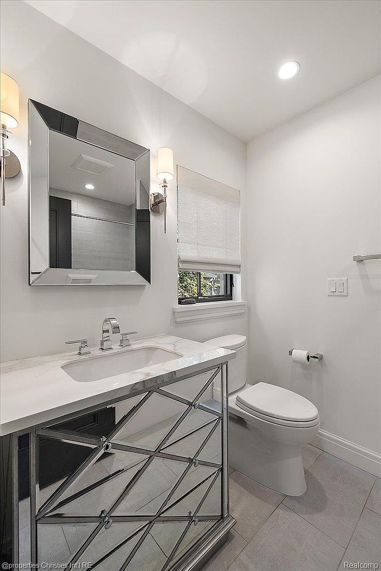This is a well-lit bathroom featuring a modern vanity with a marble countertop and a mirrored geometric design on the cabinet. A large, square mirror hangs above the sink, flanked by sconce lighting. A toilet is positioned to the right, and the floor is tiled in a neutral tone, creating a clean and contemporary aesthetic.