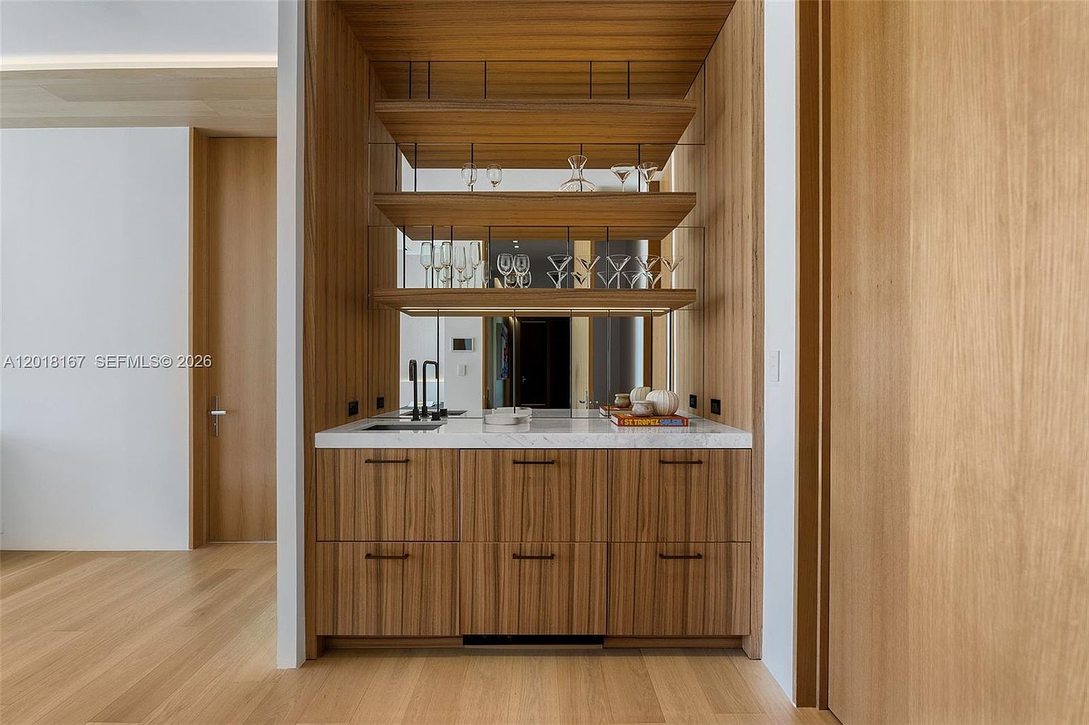 This image showcases a sophisticated built-in wet bar area featuring warm-toned wood cabinetry with a vertical grain pattern. The space includes a stone countertop, a sleek sink, and three levels of open shelving backed by a mirror, which creates a sense of depth and displays glassware. The design is clean and modern, integrated seamlessly into the surrounding wood-paneled walls and light-toned flooring.