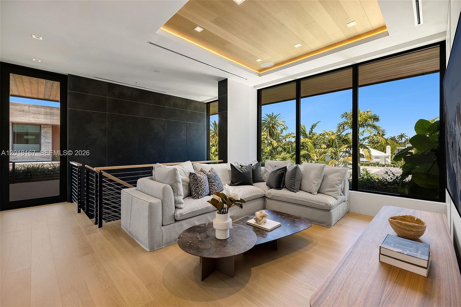 This modern living room features a light-toned wood floor and a recessed ceiling with integrated lighting. A large, light-colored sectional sofa is positioned in front of expansive windows that offer a view of lush palm trees outside. The space is accented by a dark, textured wall panel, a tiered coffee table, and a wooden console table, creating a sophisticated and airy atmosphere.