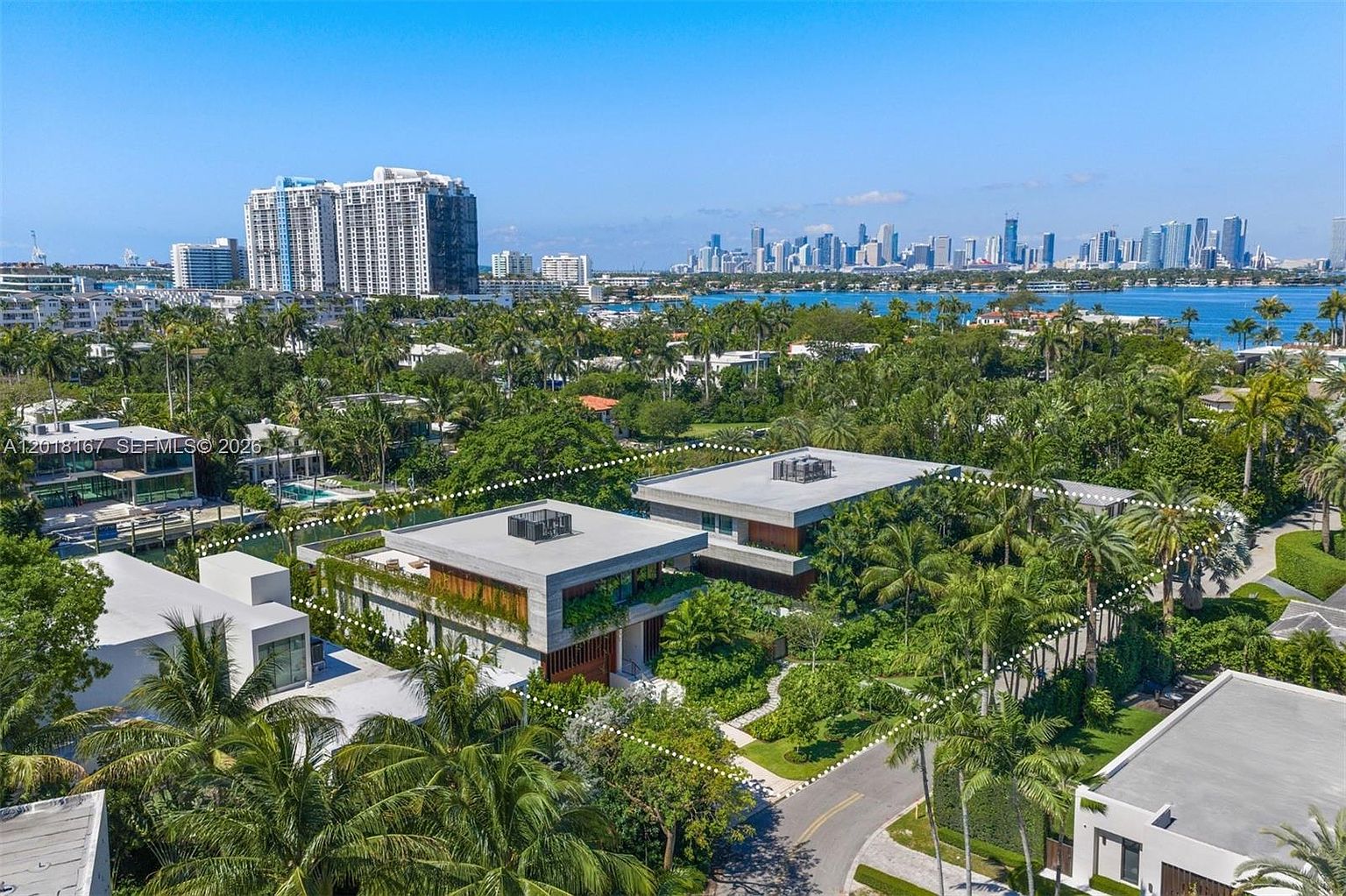 This high-angle aerial view captures a modern, multi-level residential property nestled within a lush, tropical landscape. The architecture features clean, horizontal lines, flat roofs, and a blend of concrete and wood-toned accents, surrounded by dense palm trees and manicured greenery. In the background, a serene waterway separates the property from a distant city skyline, creating a sophisticated and tranquil coastal atmosphere.