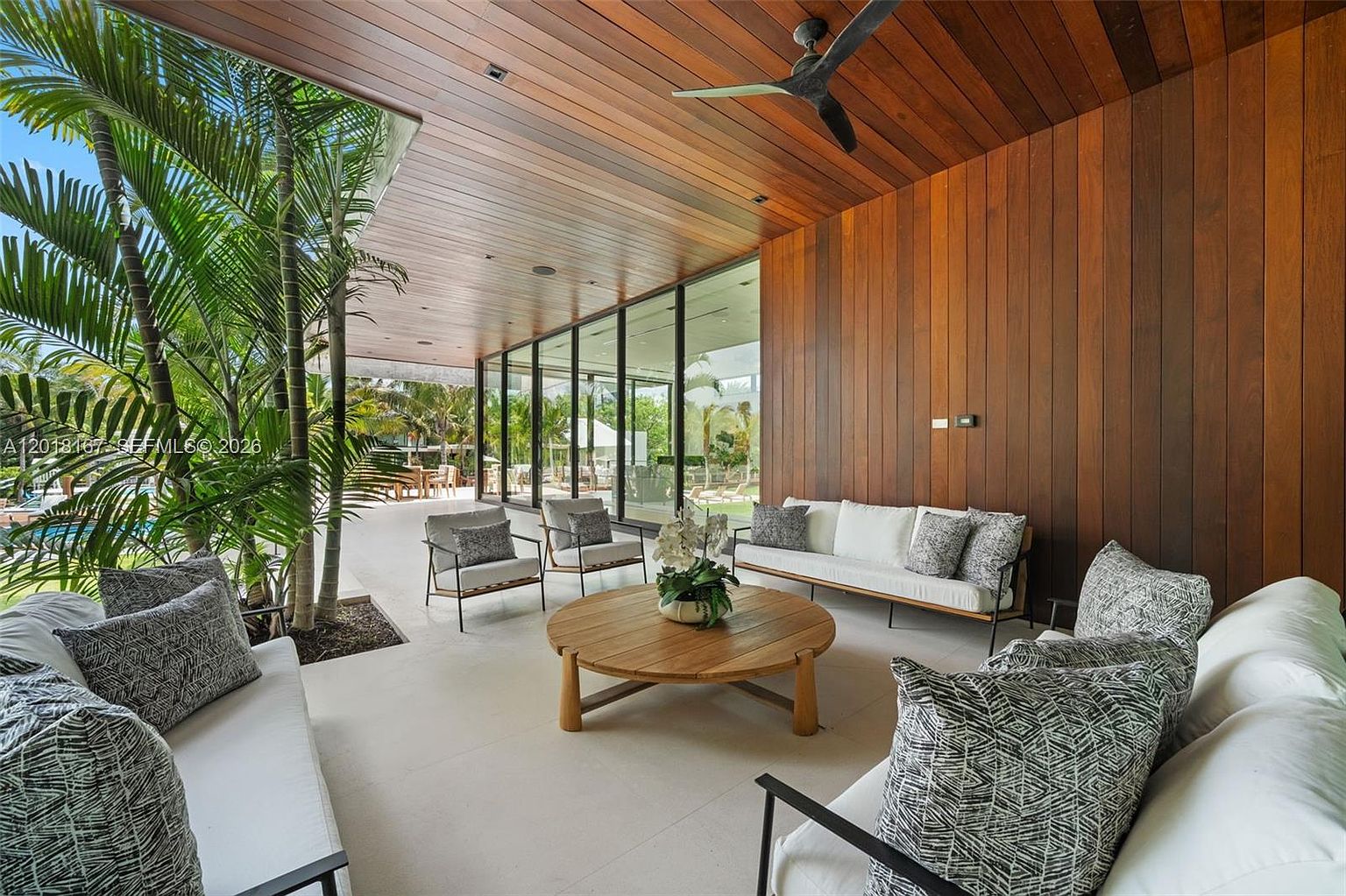 This luxurious covered patio features a warm wood-paneled ceiling and a wall of floor-to-ceiling glass that connects the indoor and outdoor living spaces. The area is furnished with a comfortable sofa set and a central round coffee table, all set against a backdrop of lush palm trees and a view of the pool area. The space offers a seamless blend of modern architectural design and tropical outdoor living.