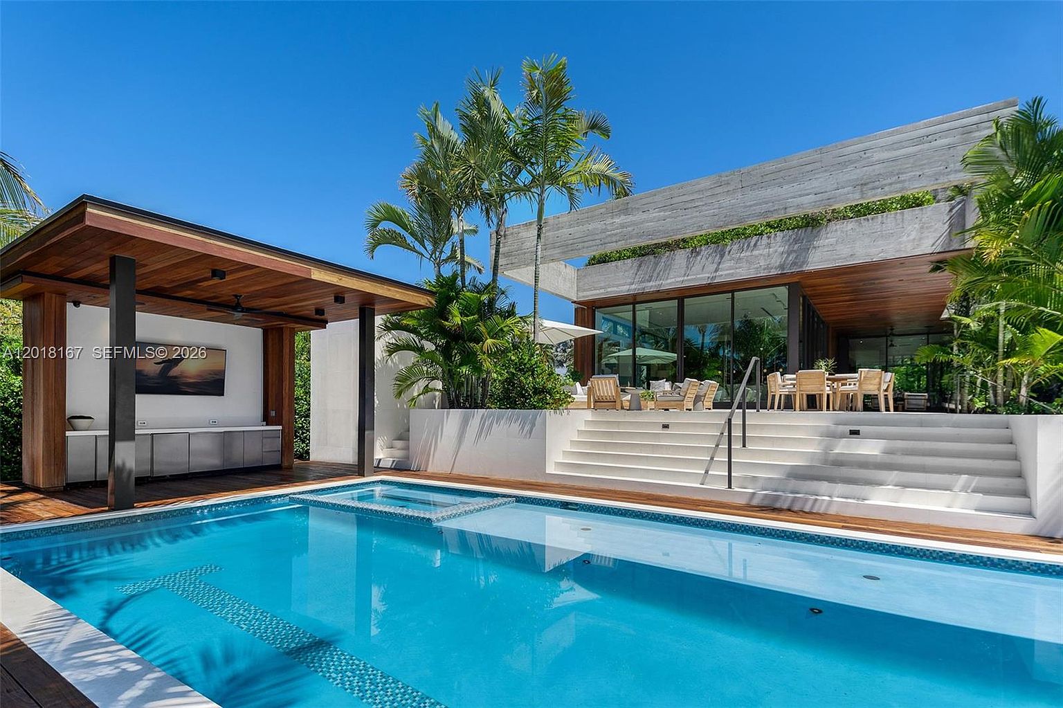 This luxurious outdoor space features a pristine rectangular swimming pool with an integrated spa, surrounded by a warm-toned wood deck. A covered outdoor kitchen area with a wall-mounted television sits to the left, while a set of wide white stairs leads up to a modern, concrete-accented patio area furnished with dining and lounge seating. The scene is framed by lush tropical palm trees and large glass walls, creating a seamless transition between the indoor and outdoor living environments.