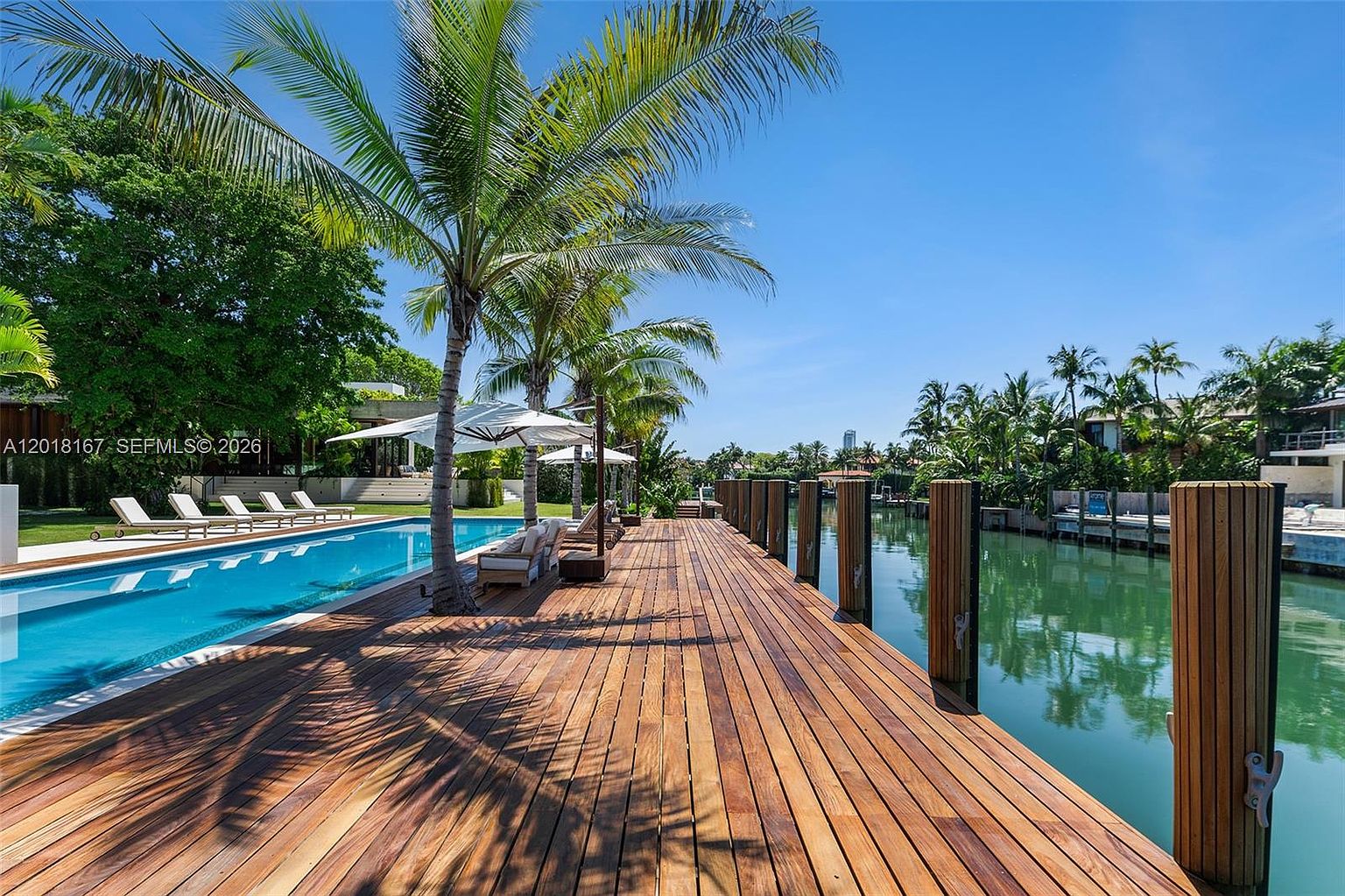 This expansive outdoor space features a long, rectangular swimming pool bordered by a wide wooden deck that extends toward a calm canal. Tall palm trees provide shade and a tropical aesthetic, while lounge chairs are arranged neatly along the pool edge. The perspective captures the seamless transition between the private waterfront deck and the serene waterway, emphasizing a luxurious, resort-like atmosphere.