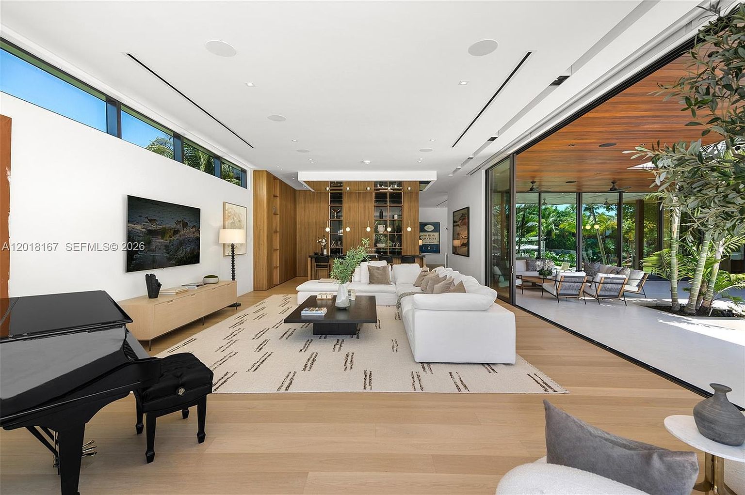 This expansive, modern living room features light wood flooring and a neutral color palette, anchored by a large white sectional sofa and a patterned area rug. A grand piano sits in the foreground, while a wall-mounted television and a sleek media console occupy the opposite wall. The space seamlessly transitions to an outdoor patio through expansive glass sliding doors, creating an airy, indoor-outdoor living experience.