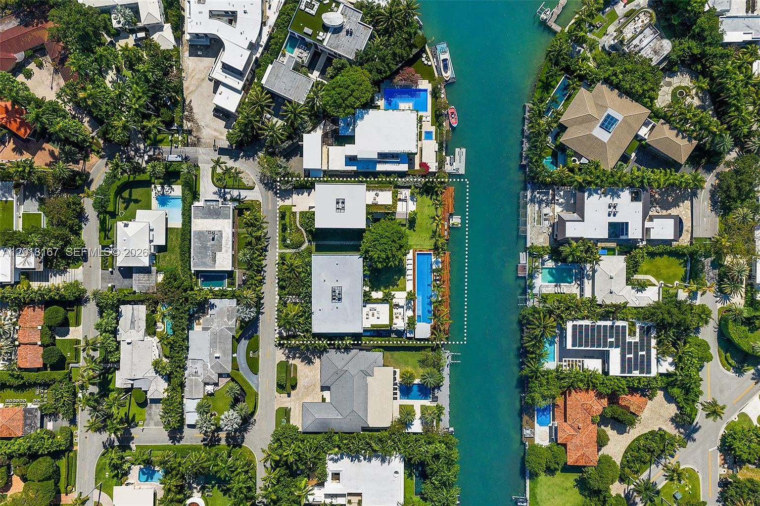 This high-altitude aerial view captures a luxurious waterfront property situated along a narrow canal in a dense, tropical residential neighborhood. The highlighted lot features a modern multi-structure residence, a long rectangular swimming pool, and lush landscaping, all bordered by a private dock. The perspective provides a comprehensive look at the property's layout, its relationship to the surrounding homes, and the direct water access.