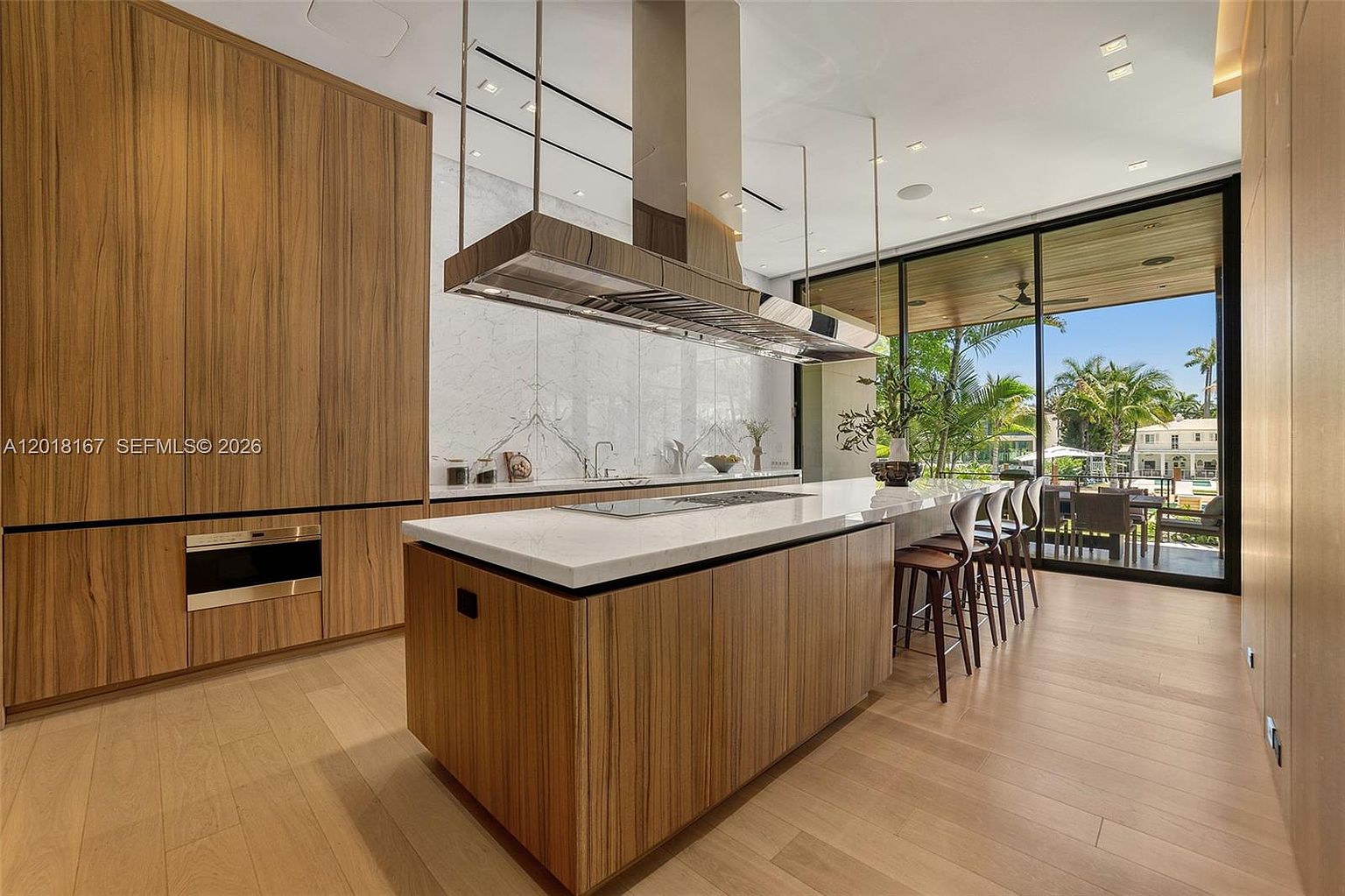 This modern, high-end kitchen features sleek, light-toned wood cabinetry and a large central island topped with a white stone surface. A prominent stainless steel range hood hangs above the island, while floor-to-ceiling glass walls provide a seamless transition to an outdoor patio area. The space is characterized by clean lines, minimalist design, and an abundance of natural light, creating an airy and sophisticated atmosphere.