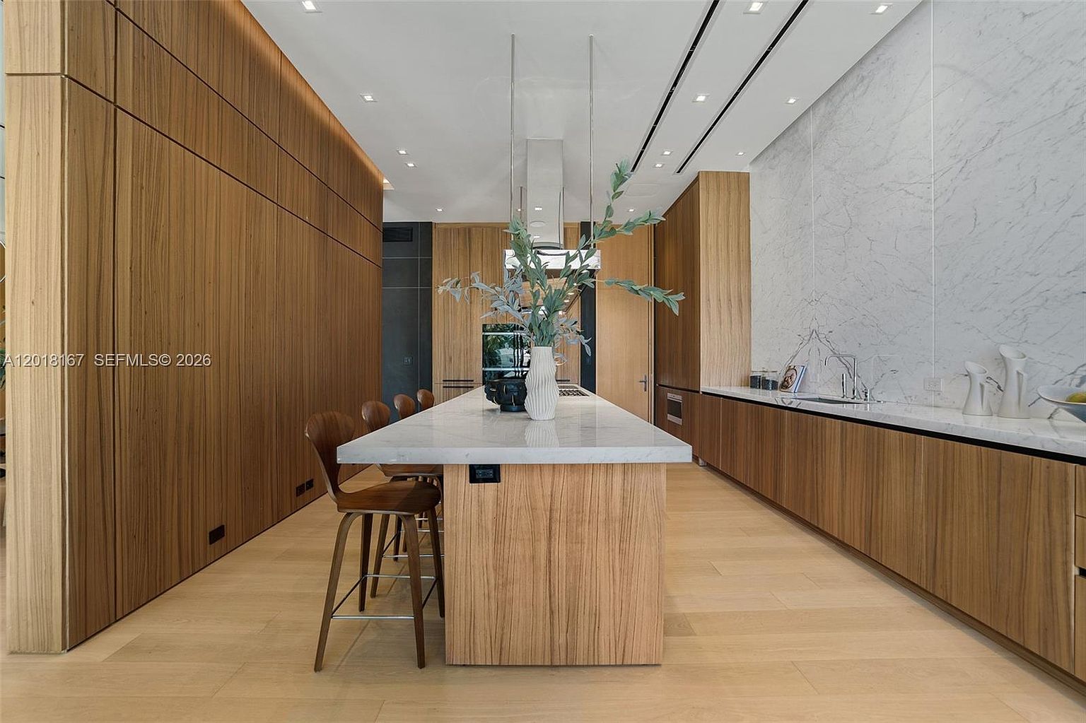 This modern, minimalist kitchen features a long central island with a stone countertop and light wood cabinetry, complemented by matching wood-paneled walls. A large, light-colored stone slab backsplash spans the wall behind the sink, creating a clean and sophisticated aesthetic. The perspective is centered, showcasing the symmetrical layout of the cabinetry and the sleek, integrated appliances.