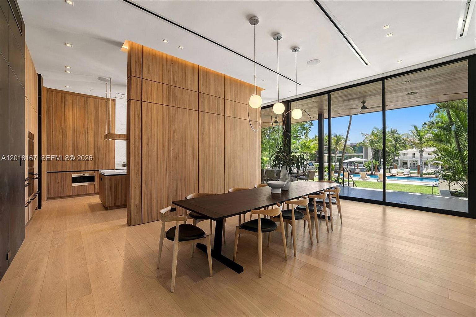 This sophisticated dining area features a long, dark wood table surrounded by modern chairs, set against a backdrop of warm, vertical wood paneling. To the right, expansive floor-to-ceiling glass walls provide a seamless transition to an outdoor pool area, bathing the room in natural light. The open-concept design flows into a sleek kitchen space, creating an airy and contemporary atmosphere perfect for entertaining.