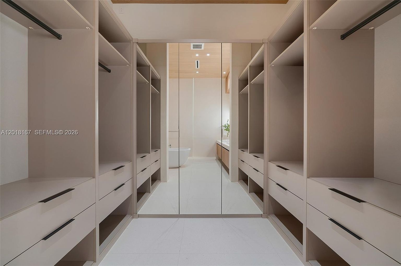 This spacious walk-in closet features custom light-toned cabinetry with ample shelving and drawer storage on both sides. A central mirrored door reflects the adjacent bathroom, which contains a freestanding tub and a vanity, creating a seamless and open transition between the two spaces. The minimalist design and neutral color palette provide a clean, organized, and luxurious aesthetic.