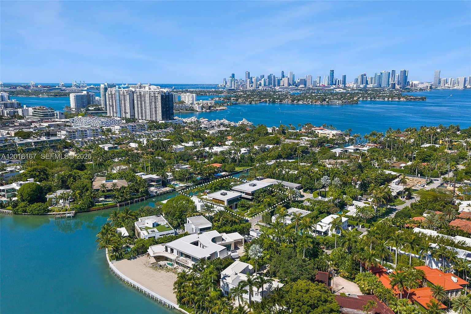 This high-angle aerial view captures a luxurious waterfront estate nestled within a lush, tropical neighborhood. The property features a modern, multi-level residence with a private dock and sandy beach access, surrounded by dense palm trees and manicured landscaping. In the background, the expansive blue bay leads to a prominent city skyline, emphasizing the prime coastal location and serene atmosphere.