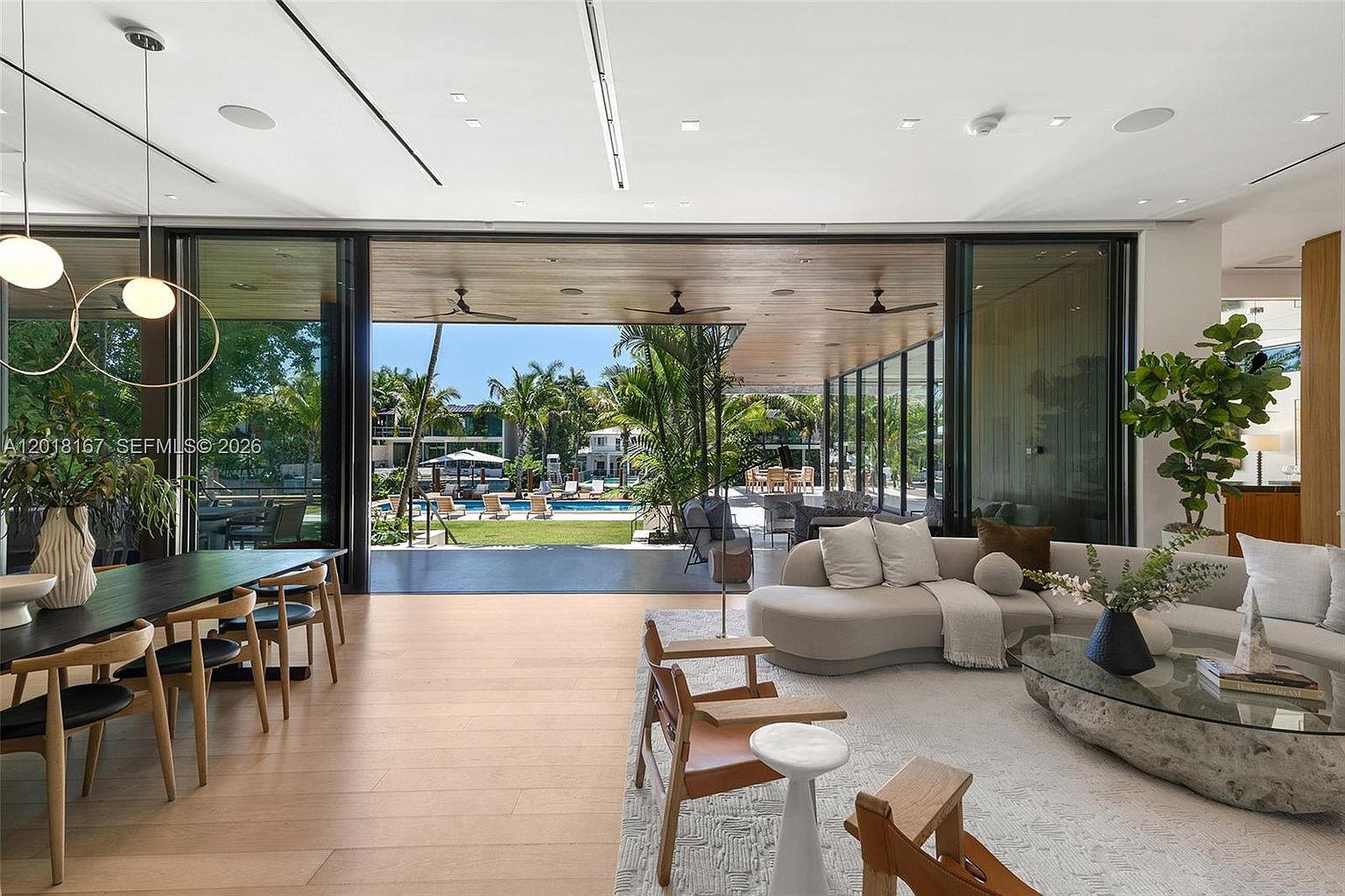 This expansive, open-concept living and dining area features a seamless transition to an outdoor patio through massive, retractable glass walls. The interior is furnished with a curved, light-toned sectional sofa, a unique stone-base coffee table, and a contemporary dining set, all set against light wood flooring. The space offers a bright, airy atmosphere with a direct, unobstructed view of a lush backyard and swimming pool, emphasizing a luxurious indoor-outdoor lifestyle.