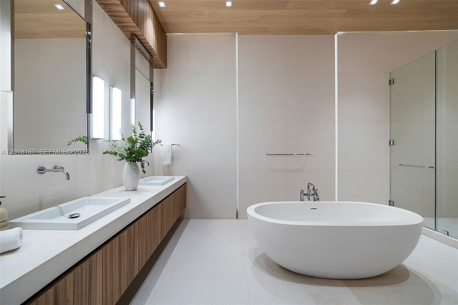 This modern primary bathroom features a sleek, minimalist aesthetic with a floating double vanity topped with light-toned stone and a large, oval freestanding soaking tub. The space is accented by warm wood-toned cabinetry and ceiling paneling, contrasting with the neutral, large-format wall and floor tiles. A glass-enclosed shower is visible to the right, and the room is illuminated by recessed lighting and vertical wall-mounted fixtures flanking the mirrors.