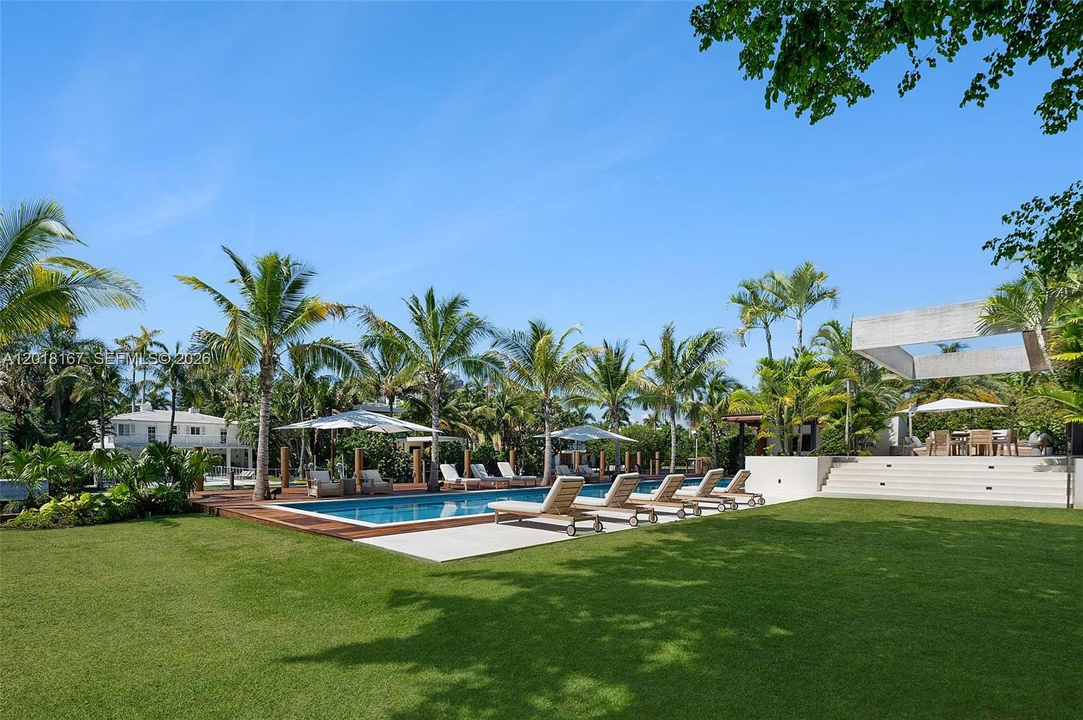 This expansive backyard features a pristine rectangular swimming pool surrounded by a wooden deck and a lush, manicured lawn. Multiple lounge chairs are arranged poolside, complemented by white umbrellas and a raised patio area with outdoor dining furniture under a modern architectural structure. The scene is framed by mature palm trees and dense greenery, creating a private and serene tropical oasis.