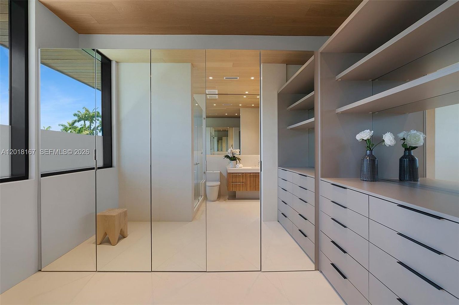 This modern walk-in closet features a clean, minimalist aesthetic with light-toned cabinetry and integrated shelving. A series of mirrored doors reflects the adjacent bathroom, creating a sense of depth and continuity between the spaces. The room is finished with a light wood ceiling and neutral flooring, providing a bright and organized atmosphere.