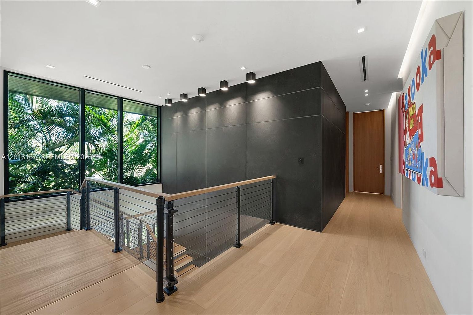 This modern hallway and staircase area features light wood flooring and a striking dark, matte-finished accent wall illuminated by a row of sleek, square ceiling spotlights. A staircase with a metal railing and wood handrail descends alongside large windows that provide a view of lush tropical greenery outside. The space is further accented by a large, colorful pop-art style wall hanging, creating a sophisticated and contemporary aesthetic.